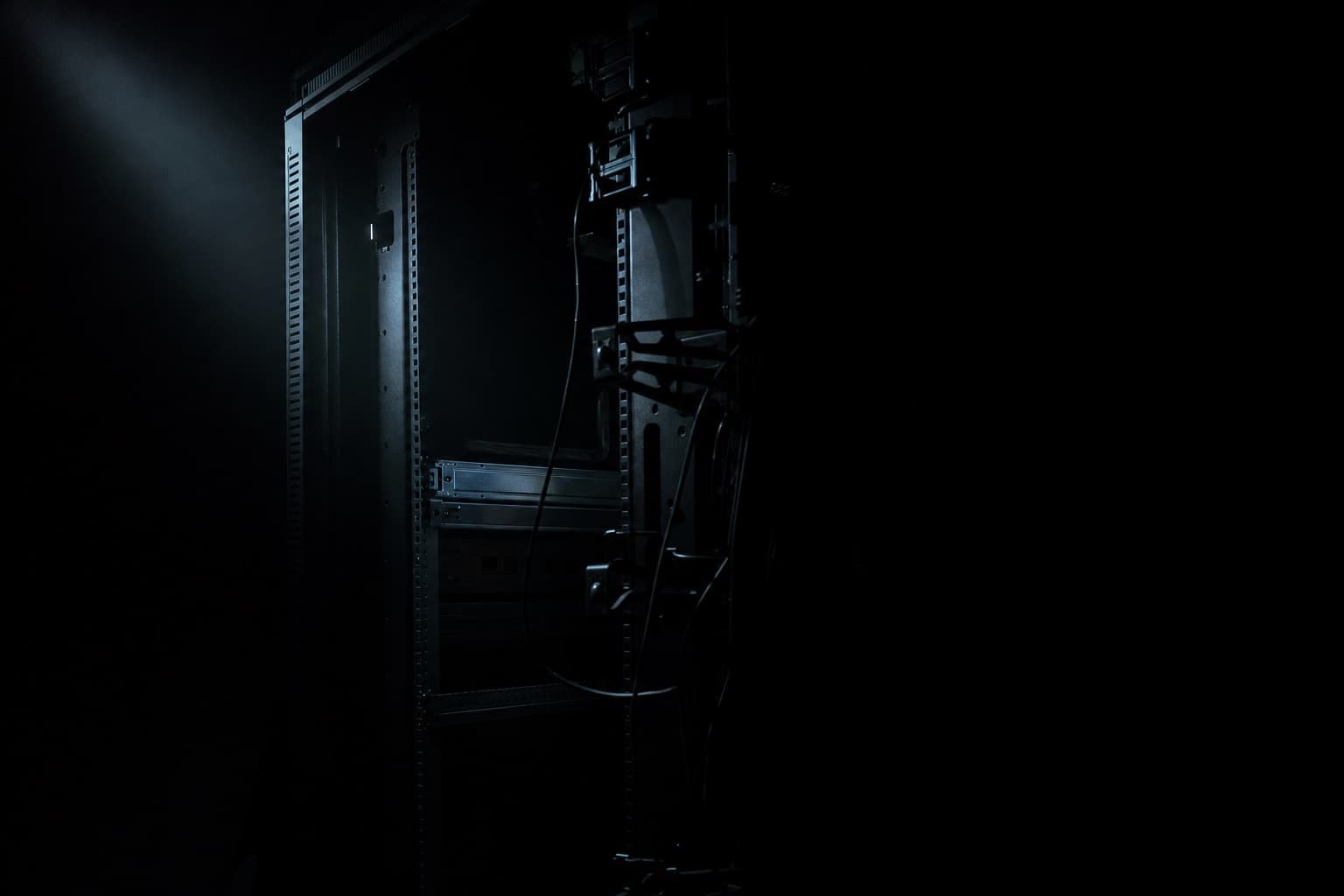 The rear of a half-emptied server rack lit by a single narrow beam, its cable management arms hanging loose